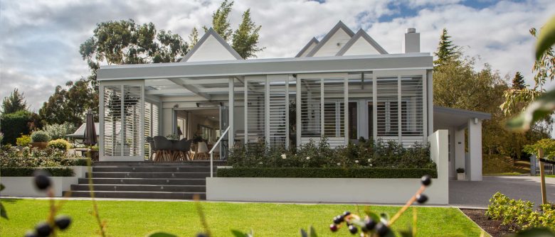 View across a lush green lawn to the front of the house, featuring sliding shutters on all sides of an enclosed porch, blending modern design with natural surroundings.