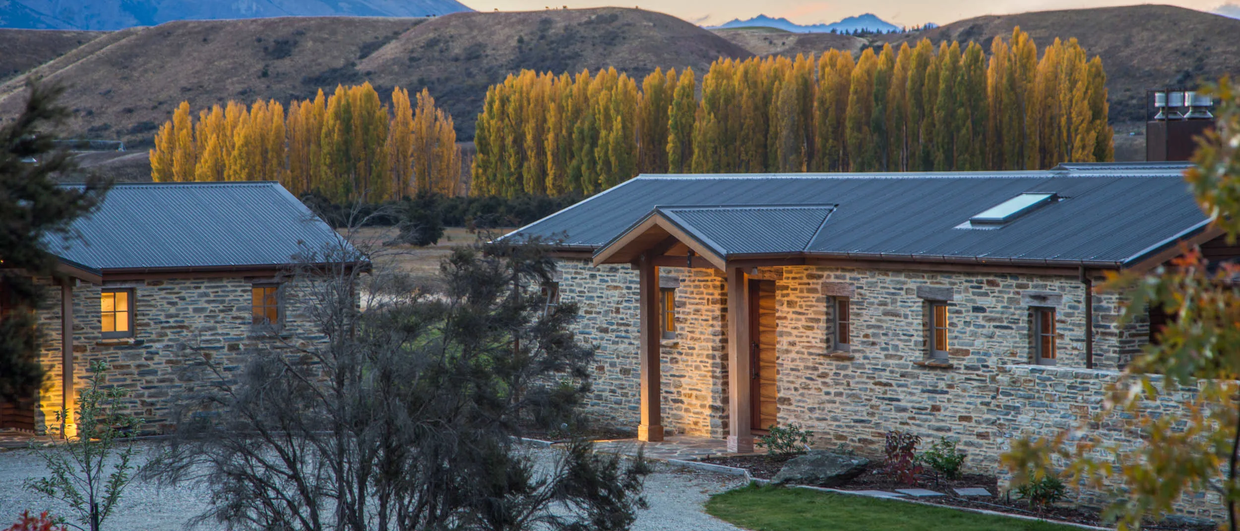View to the entry of the schist clad Stevenson Rd house and cottage with golden poplars behind house in an early Autumn evening.