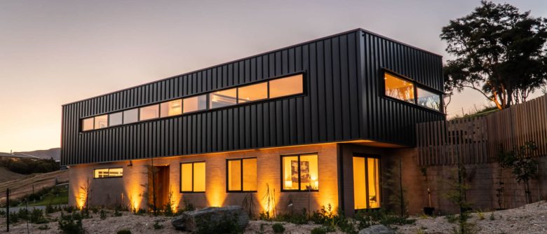 Dusk street view highlighting the home’s pale brick and dark long-run iron cladding, with the warm glow of interior lighting.