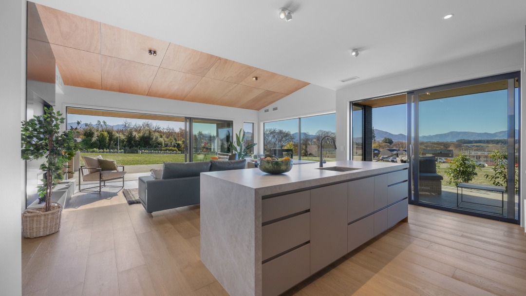 View across the kitchen toward the open-plan living area, highlighting seamless flow, natural light, and a built-in window seat framing the landscape beyond.