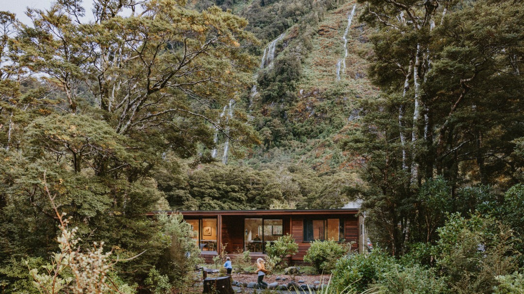 Looking towards chalets set against a dramatic backdrop of native bush and a waterfall, showcasing a serene natural alpine setting.