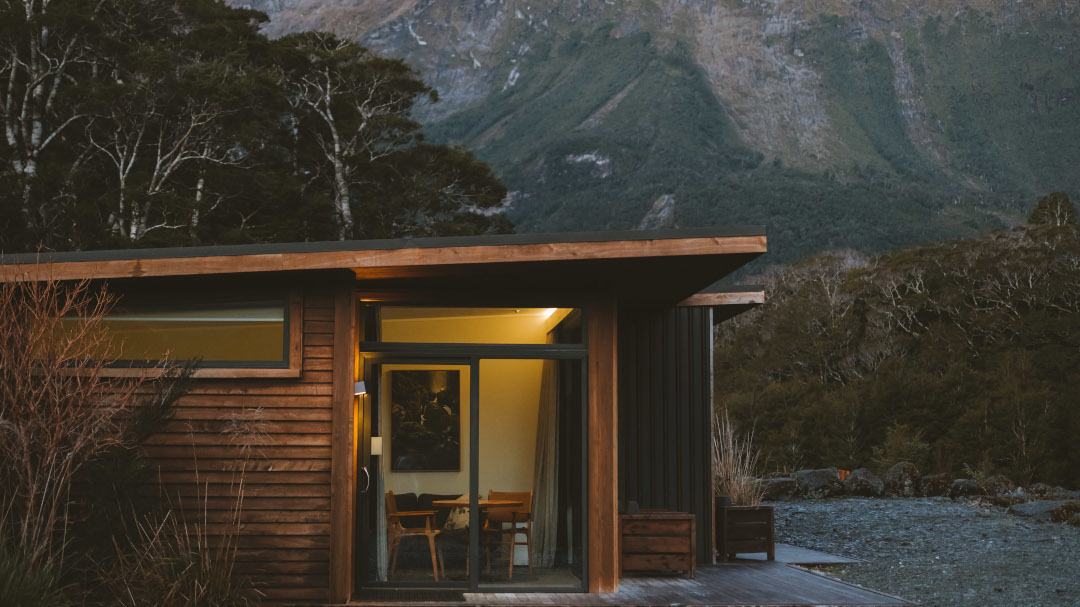 Side view at dusk of a luxury Milford Sound Lodge chalet set against a dramatic mountain backdrop, with soft evening light and native bush.