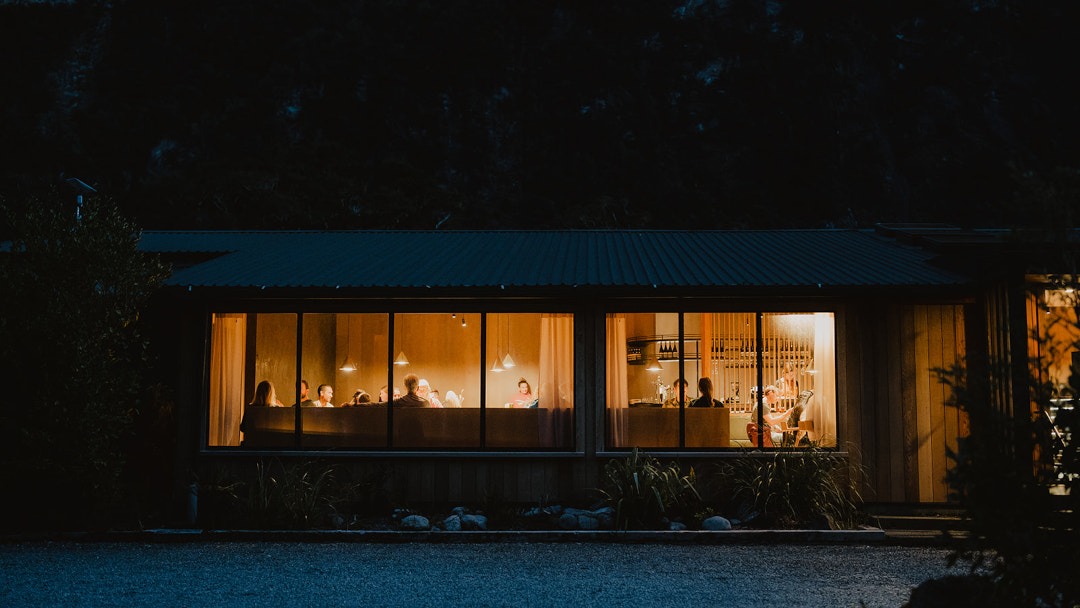 From the outside looking into the warm interior of the restaurant, lounge, and bar areas at night, with glowing lights and inviting timber-lined spaces.